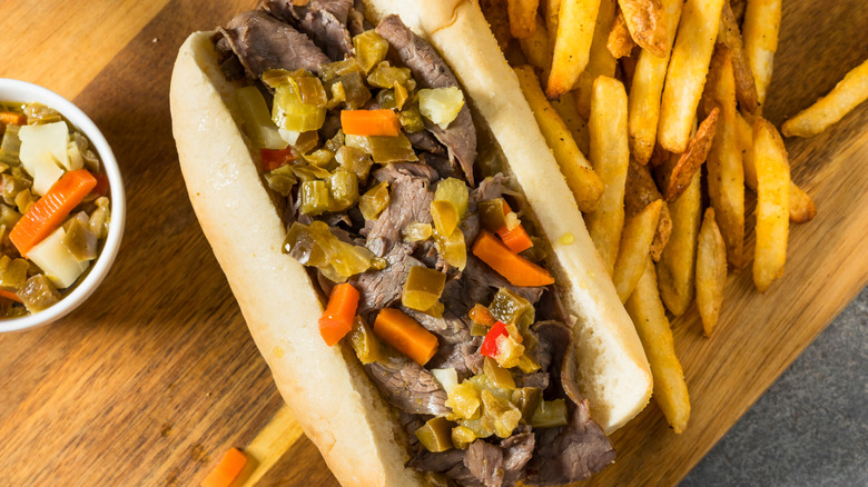 Beef sandwich with giardiniera sitting on a wood board near a pile of french fries.
