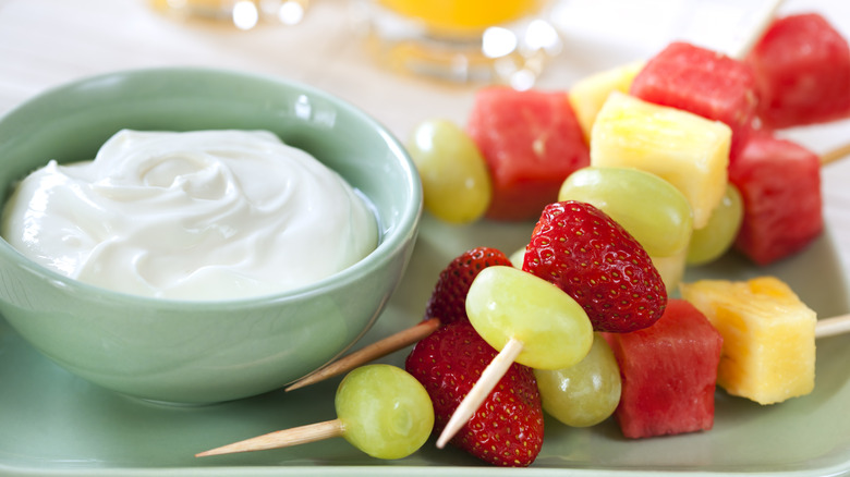 A bowl of yogurt dip next to fruit skewers