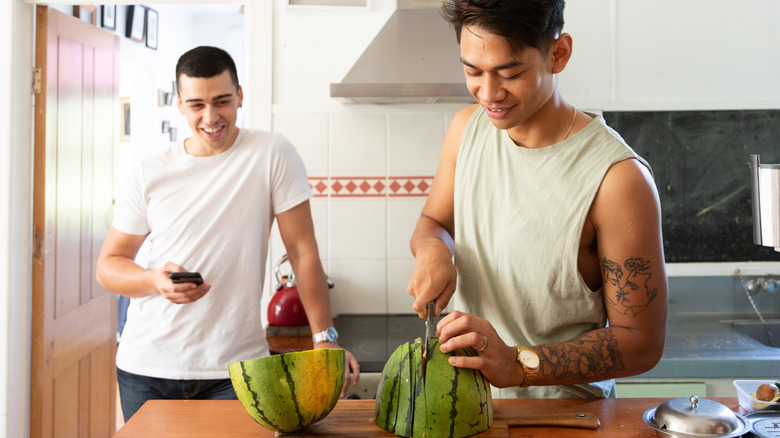 Two men in the kitchen with one slicing watermelon