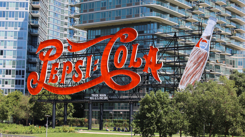 Pepsi-Cola billboard in classic cursive letters and soda bottle in park front of high-rise buildings