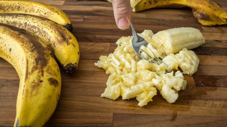 A hand mashing up a banana with a fork, surrounded by more bananas in peels