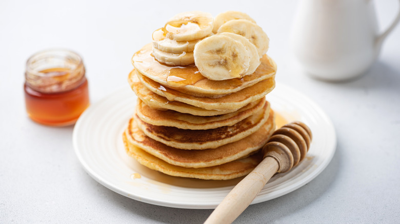 A stack of banana pancakes topped with bananas and honey with a honey-dipper on the plate