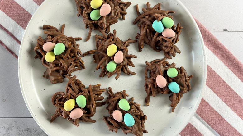 nest-shaped chocolate cookies filled with candy eggs on a white plate sitting on top of a red-and-white-striped towel