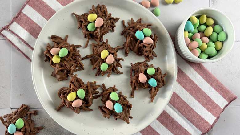 nest-shaped chocolate cookies filled with candy eggs on a white plate sitting on top of a red-and-white-striped towel