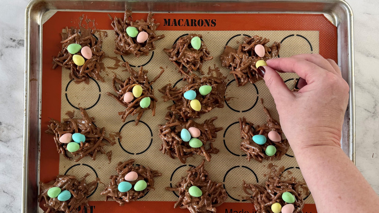 hand pinching a candy egg while holding it over a nest-shaped chocolate cookie on a baking pan