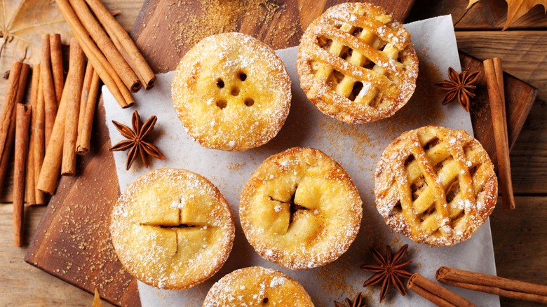 Six different mini apple pies dusted with powdered sugar next to star anise and cinnamon sticks on wooden table