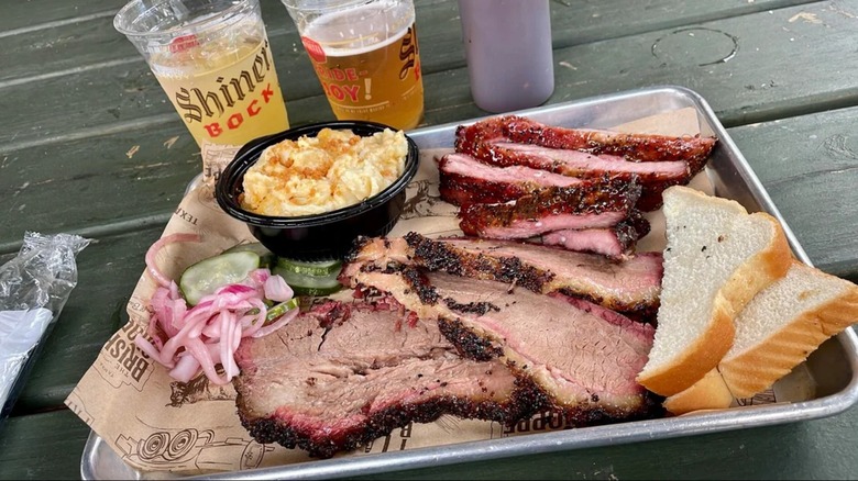 A platter featuring brisket, mac and cheese, and bread, with two drinks in plastic cups