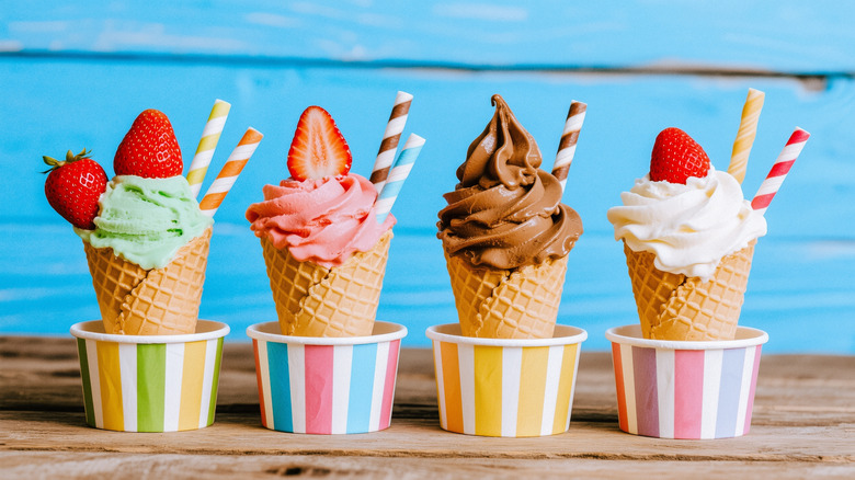 A row of ice cream cones in different flavors, standing in colorful striped paper cups with strawberries and striped straws for decoration