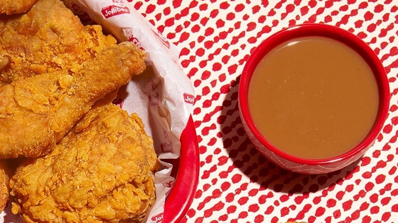 A small red bowl of gravy rests to the right of a basket of Jollibee fried chicken.