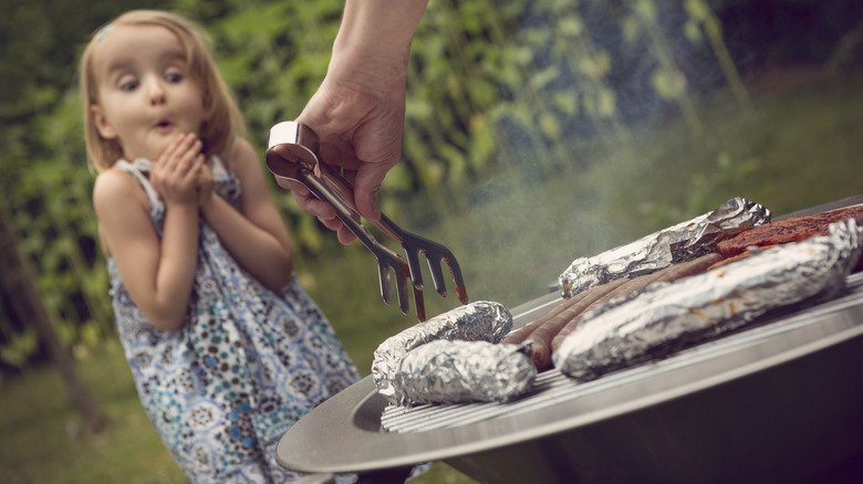 A little girl in a dress excited about hot dogs and foil-wrapped food on a BBQ