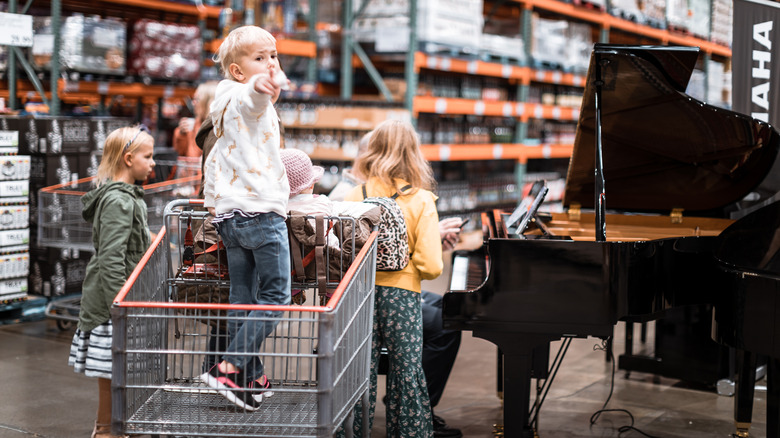 Boy standing in a Costco shopping cart and two girls standing next to it, near a piano with Costco shelves in the background