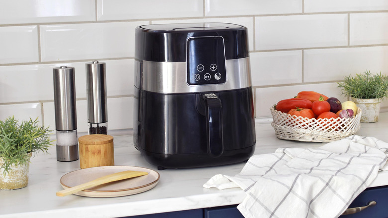 Air fryer on kitchen countertop surrounded by basket of tomatoes and salt and pepper grinders