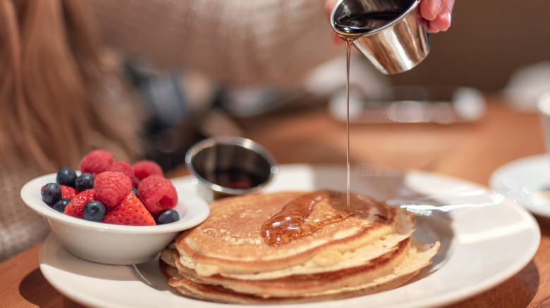 Woman pouring syrup on pancakes on a white plate with a small bowl of berries