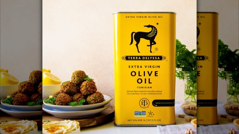 A can of Terra Delyssa Extra Virgin Olive Oil next to a plate of falafel, hummus, and fresh herbs