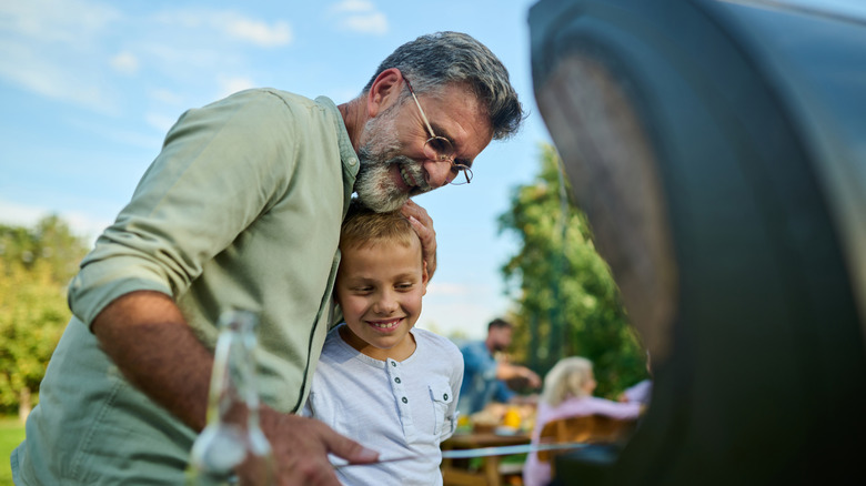 Grandfather embraces grandson while watching over the grill outside in the summer.