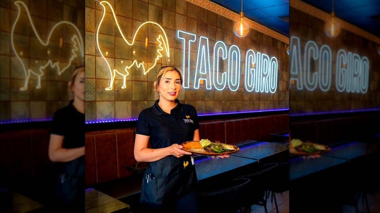 A server holds a plate of food in front of a Taco Giro neon sign