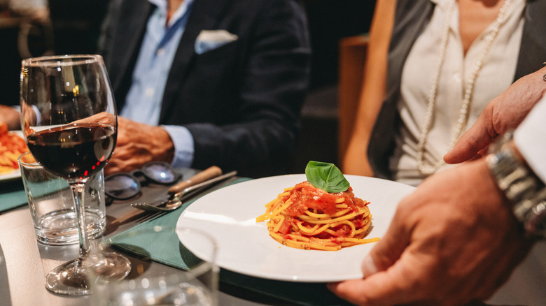 A waiter's hand places a plate with pasta in front of a diner seated at a table next to another diner.