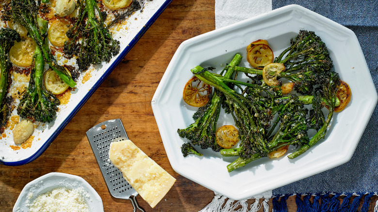 Roasted broccolini on a pan and on a white plate with lemon near a cheese grater with parmesan cheese