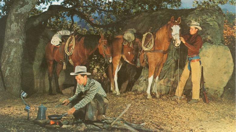 An old, staged photo of two cowboys in the morning. One is cooking food and the other is preparing the horses.