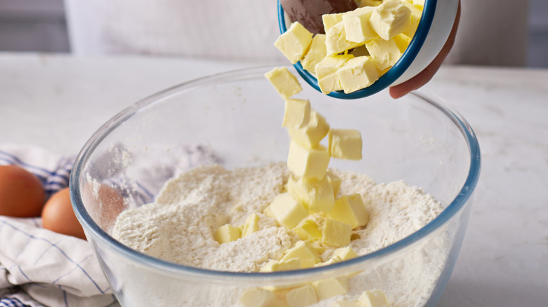 Person pouring cubed butter from a bowl into a glass bowl of flour