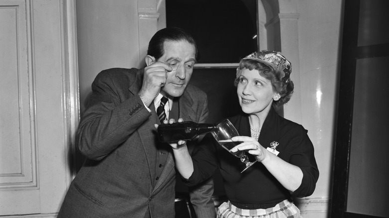 A man with a monocle watching a woman pour out a beer