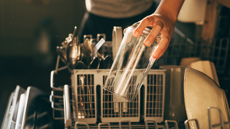 Hand holding glass cup over an opened dishwasher filled with bowls and utensils