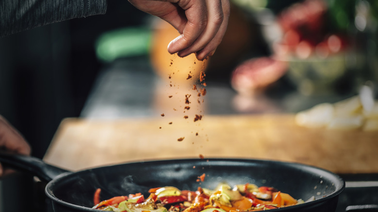 a cook sprinkles seasoning over a skillet of vegetables