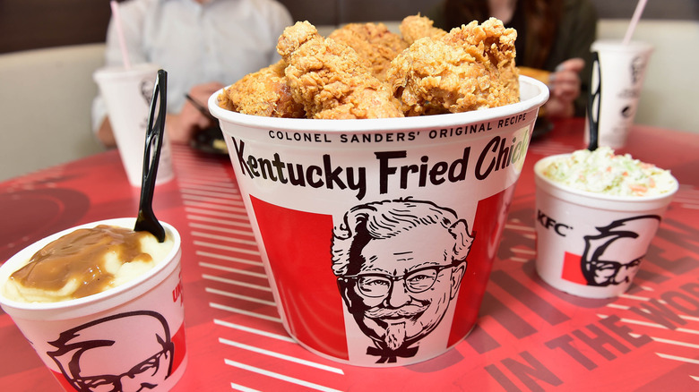 A red and white striped bucket filled with Kentucky Fried Chicken on a table next to smaller containers of mashed potatoes and gravy, and coleslaw.