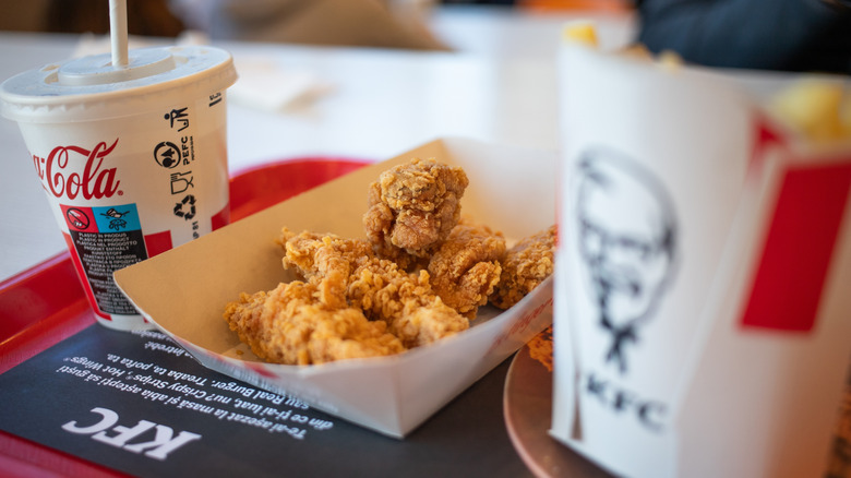 A paper boat of chicken tenders on a red plastic tray with a soda and container of fries