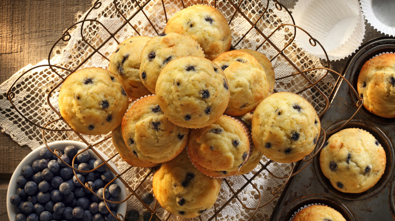 blueberry muffins stacked in a wire basket next to a muffin tin and bowl of blueberries