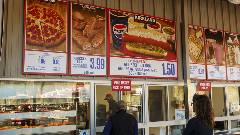 An outdoor Costco food court displaying menu items such as the cheese and pepperoni pizzas on the wall above.