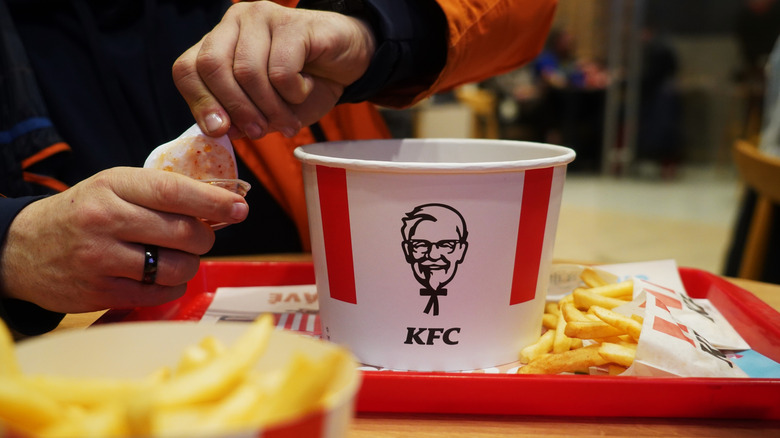 A man opening a sauce container next to a bucket of KFC chicken and some fries on a tray.