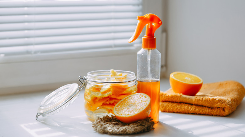 Two halves of an orange on cleaning scrubs and rags, and a container of orange peels next to a filled spray bottle