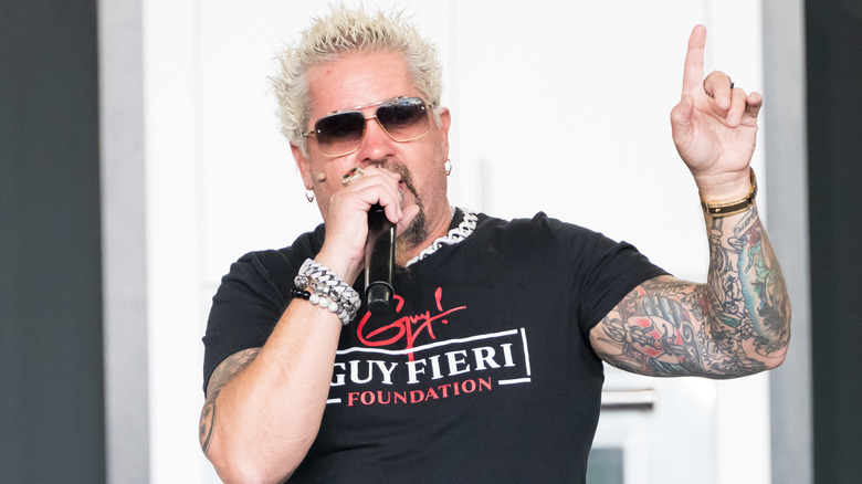 Guy Fieri wearing sunglasses and speaking into a handheld microphone at an event