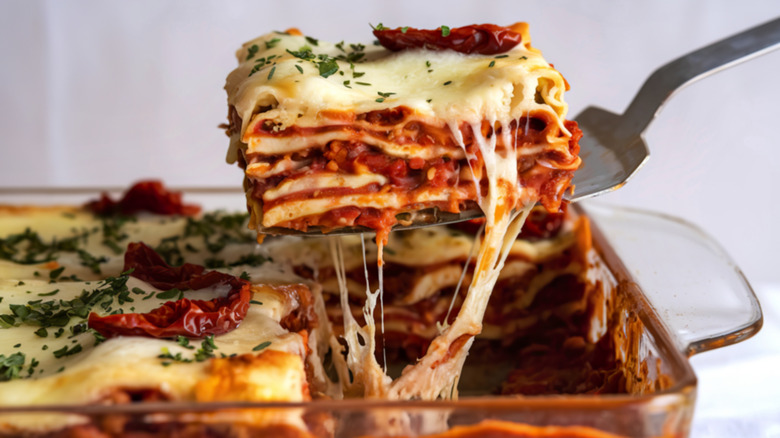 A layered lasagna topped with sun-dried tomatoes being served from a glass pan