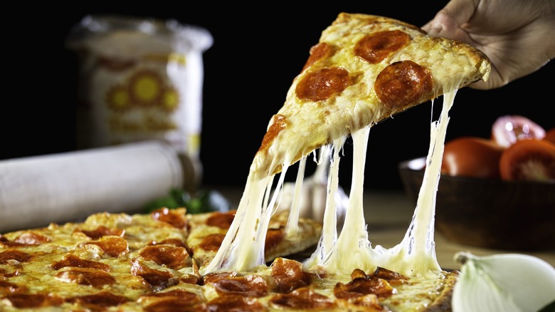 A hand lifting a slice of pepperoni pizza with a cheese pull from a whole pizza