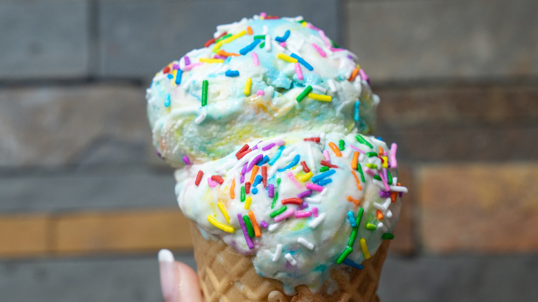 Hand holding ice cream cone with two scoops of birthday cake ice cream on top.