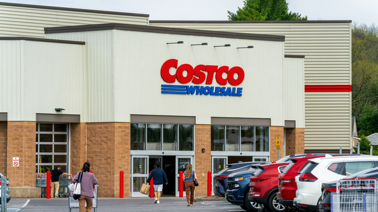 The exterior of a Costco storefront seen from the parking lot with cars and people around.