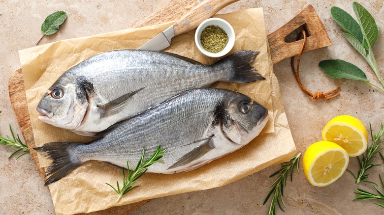 Two sea bream on a cutting board with herbs and lemons.