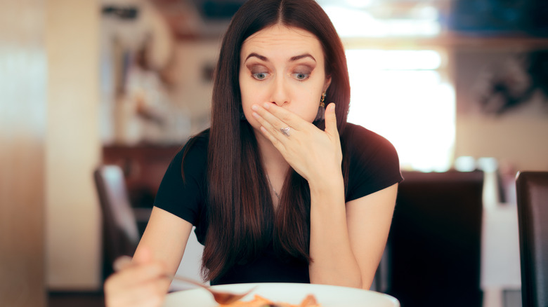 A woman eating food and holding her hand to her mouth looking surprised and nauseous.