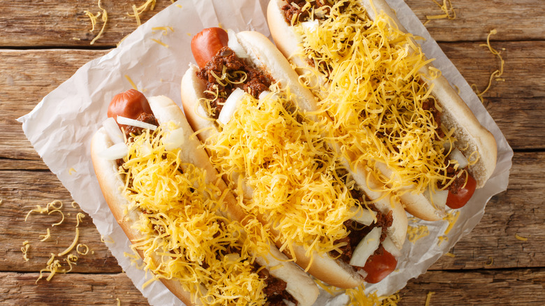 Three hot dogs in buns with chili, onion, shredded cheese