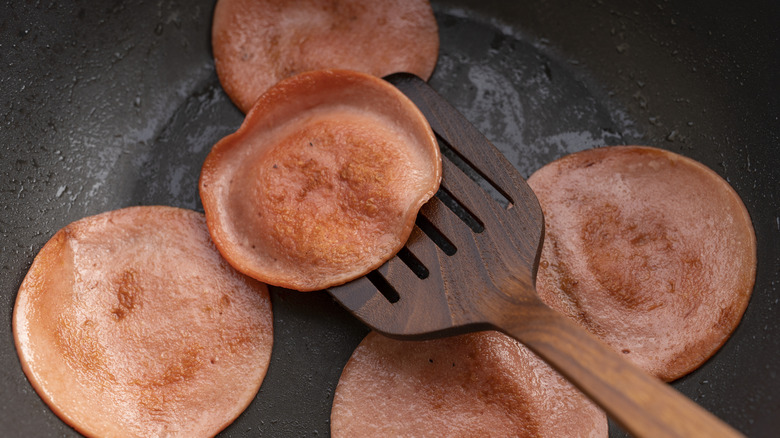 Five slices of bologna frying in a pan with one piece being lifted by a wooden spatula