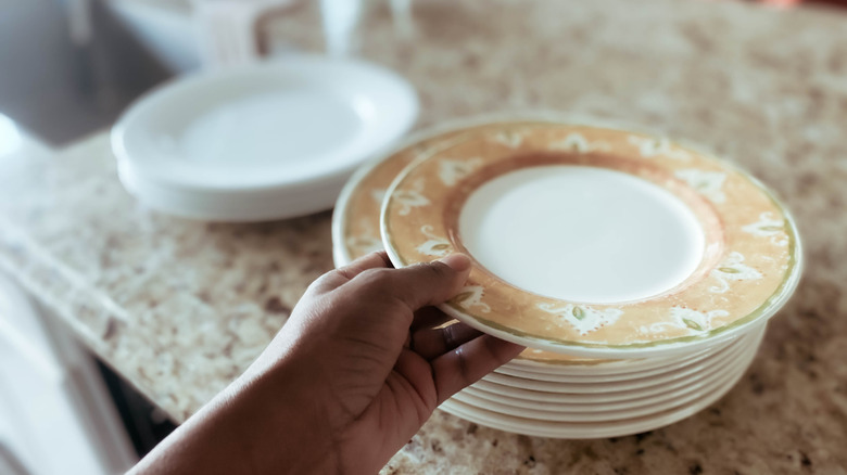 A hand stacking white and orange rimmed dishes on a granite countertop