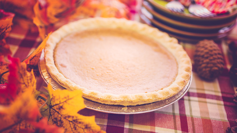 A store-bought pumpkin pie on a table with leaves, plates, and a pinecone beside it.