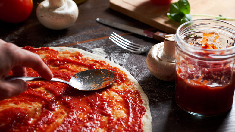 Close-up of a person spreading tomato sauce on pizza dough with other ingredients on the table in the background.