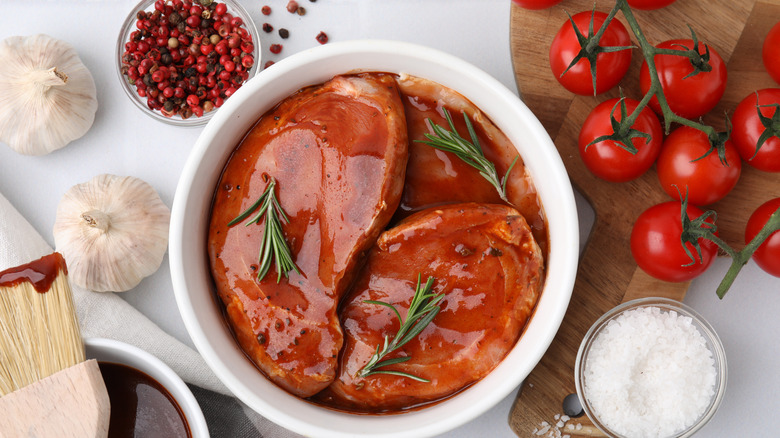 A bowl of meat marinating, surrounded by garlic, peppercorns, tomatoes, and salt.