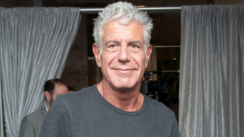 Anthony Bourdain smiling and wearing a gray T-shirt with a gray curtain in the background