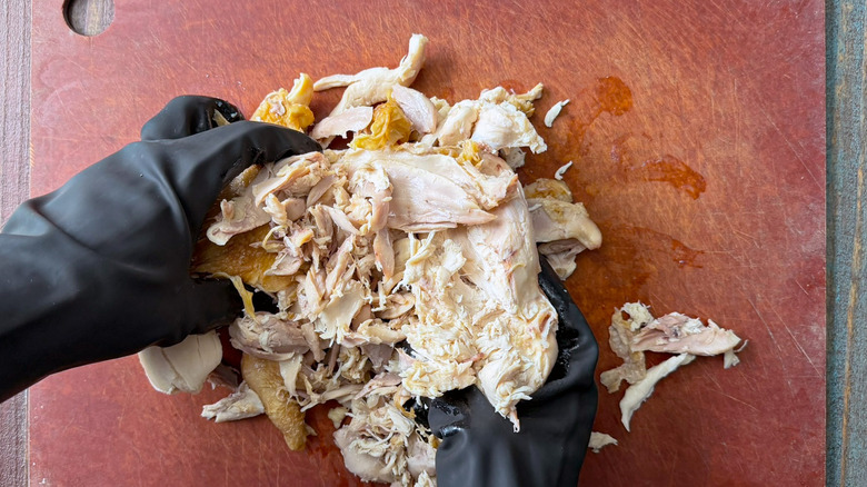 gloved hands shred a rotisserie chicken on a cutting board