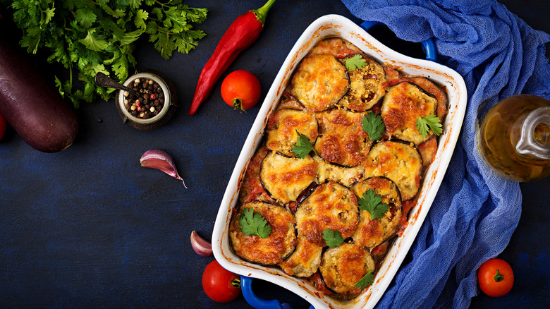 Eggplant Parmesan in a white dish on a blue surface surrounded by tomatoes, a chili pepper, a clove of garlic, a jar of spices, a bottle of oil, some celery and a raw eggplant.