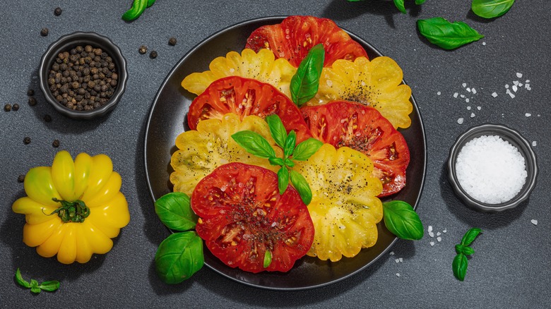 Red and yellow heirloom tomato slices on a plate with basil sprigs and a yellow tomato, basil leaves, and two small bowls of coarse salt and black peppercorns on a black surface